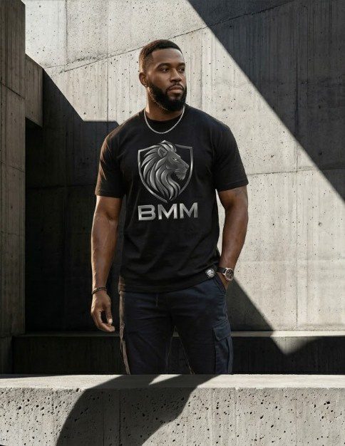 A muscular man in a premium black heavyweight t-shirt featuring a silver metallic lion head inside a shield with "BMM" printed in bold chrome letters.